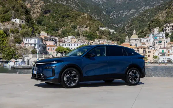 Blue Lancia Gamma electric SUV parked by a waterfront village and mountain backdrop, HD desktop wallpaper.