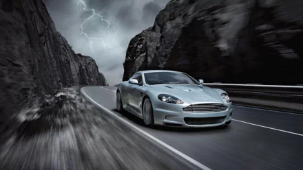 A silver Aston Martin One-77 speeding on a winding mountain road under a stormy sky, captured in an HD PC desktop wallpaper background.