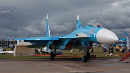 A striking HD image of a Sukhoi Su-27 military jet on the tarmac, showcasing its blue and white design against a dramatic cloud-filled sky.