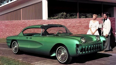 Green Chevrolet Biscayne classic parked by a mid‑century brick wall with a man and woman nearby — HD PC desktop wallpaper background, high-detail.