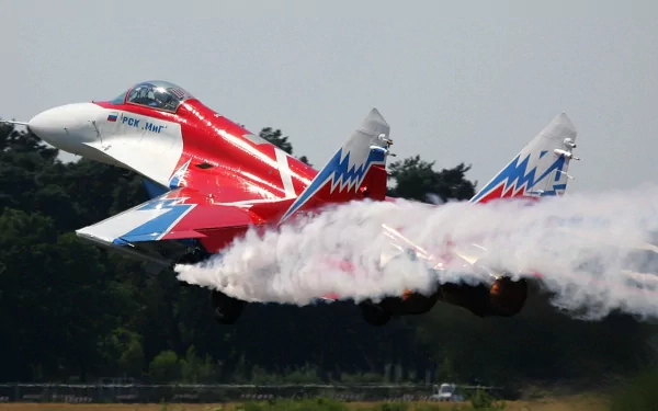 HD military-themed PC desktop wallpaper featuring a Mikoyan MiG-29 fighter jet with vibrant red, white, and blue colors, releasing smoke during flight.