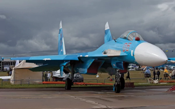 A striking HD image of a Sukhoi Su-27 military jet on the tarmac, showcasing its blue and white design against a dramatic cloud-filled sky.