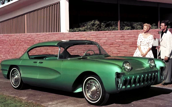 Green Chevrolet Biscayne classic parked by a mid‑century brick wall with a man and woman nearby — HD PC desktop wallpaper background, high-detail.