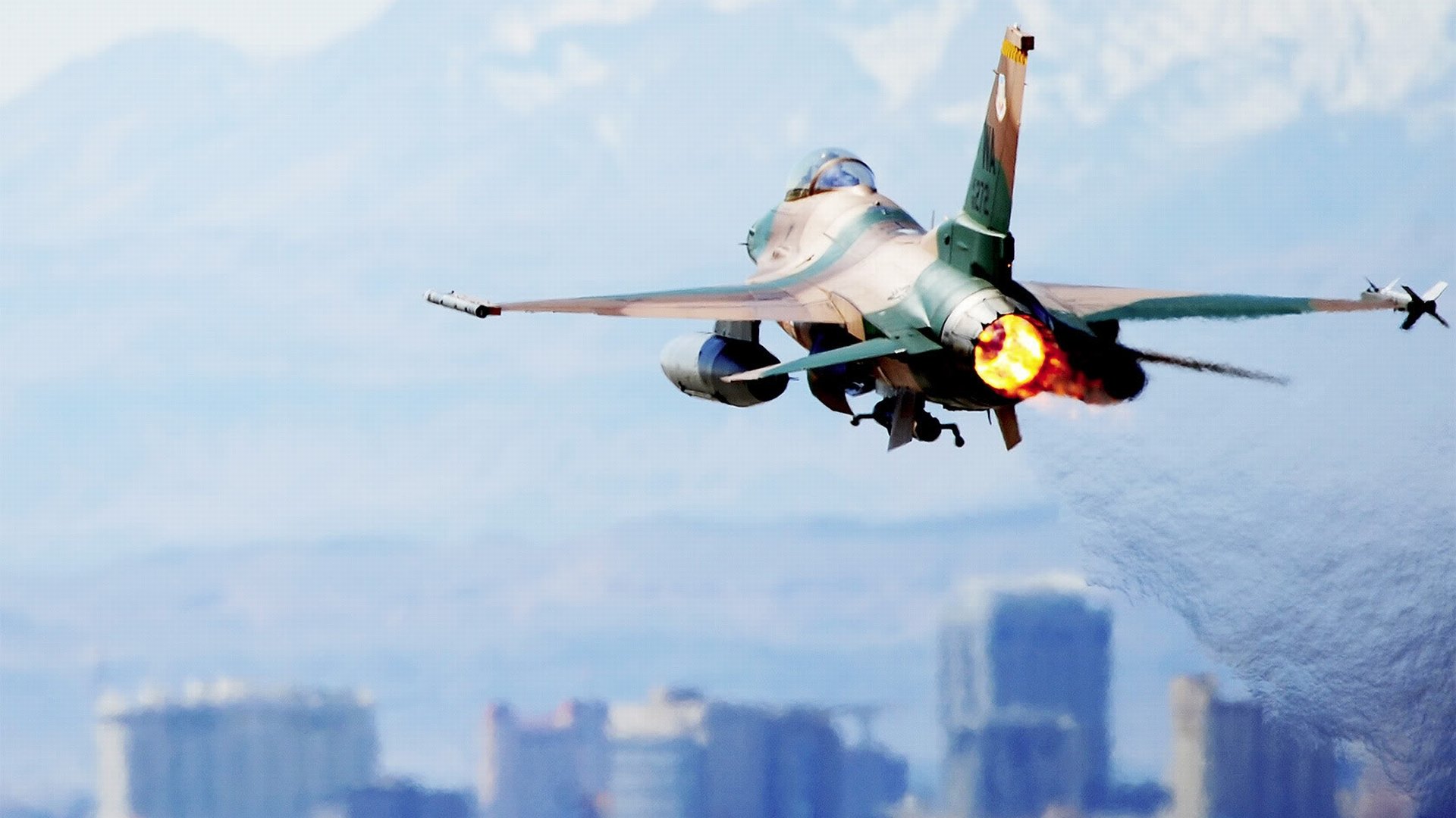 HD desktop wallpaper showcasing a General Dynamics F-16 Fighting Falcon fighter jet in flight with afterburner engaged above a blurred cityscape.
