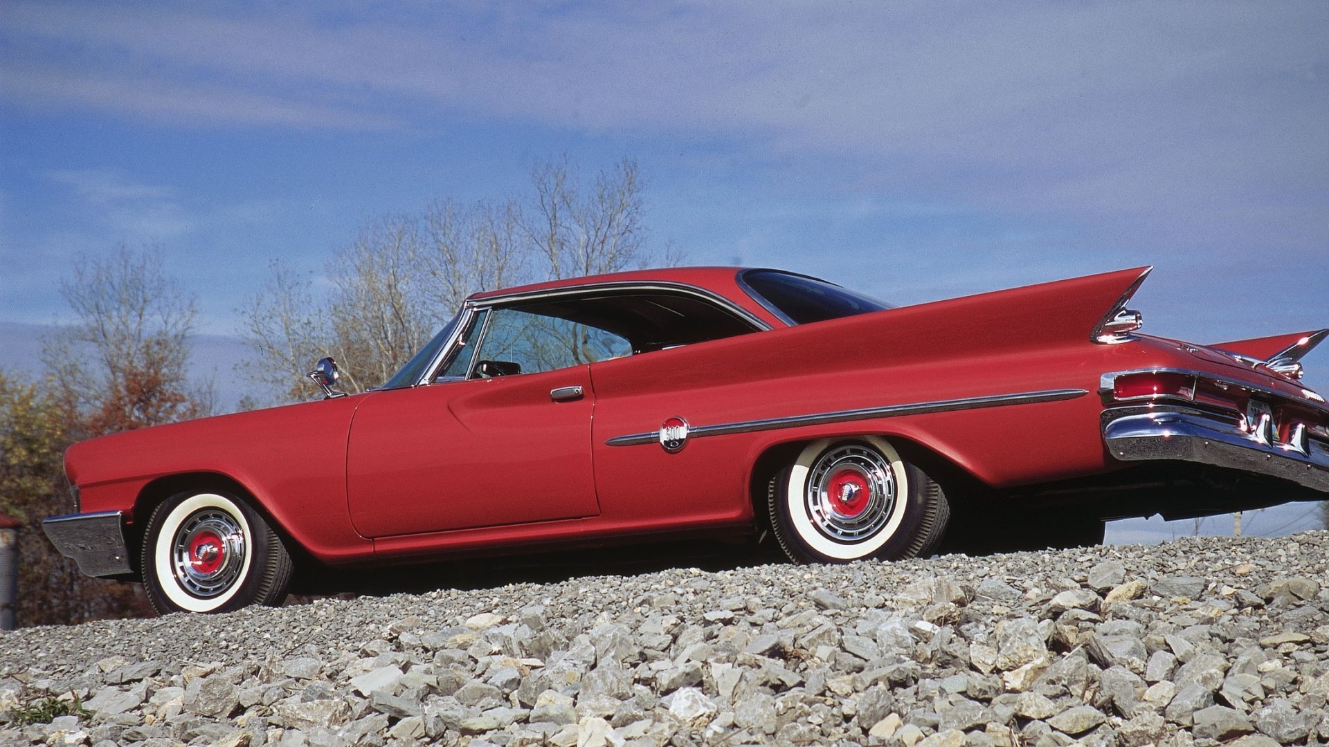 HD PC desktop wallpaper featuring a red Chrysler vehicle in low side profile, parked on gravel under a clear blue sky.