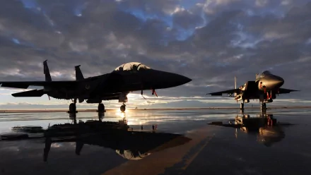 HD desktop wallpaper featuring two McDonnell Douglas F-15 Eagle military fighter jets on a reflective runway at sunset under a dramatic cloudy sky.