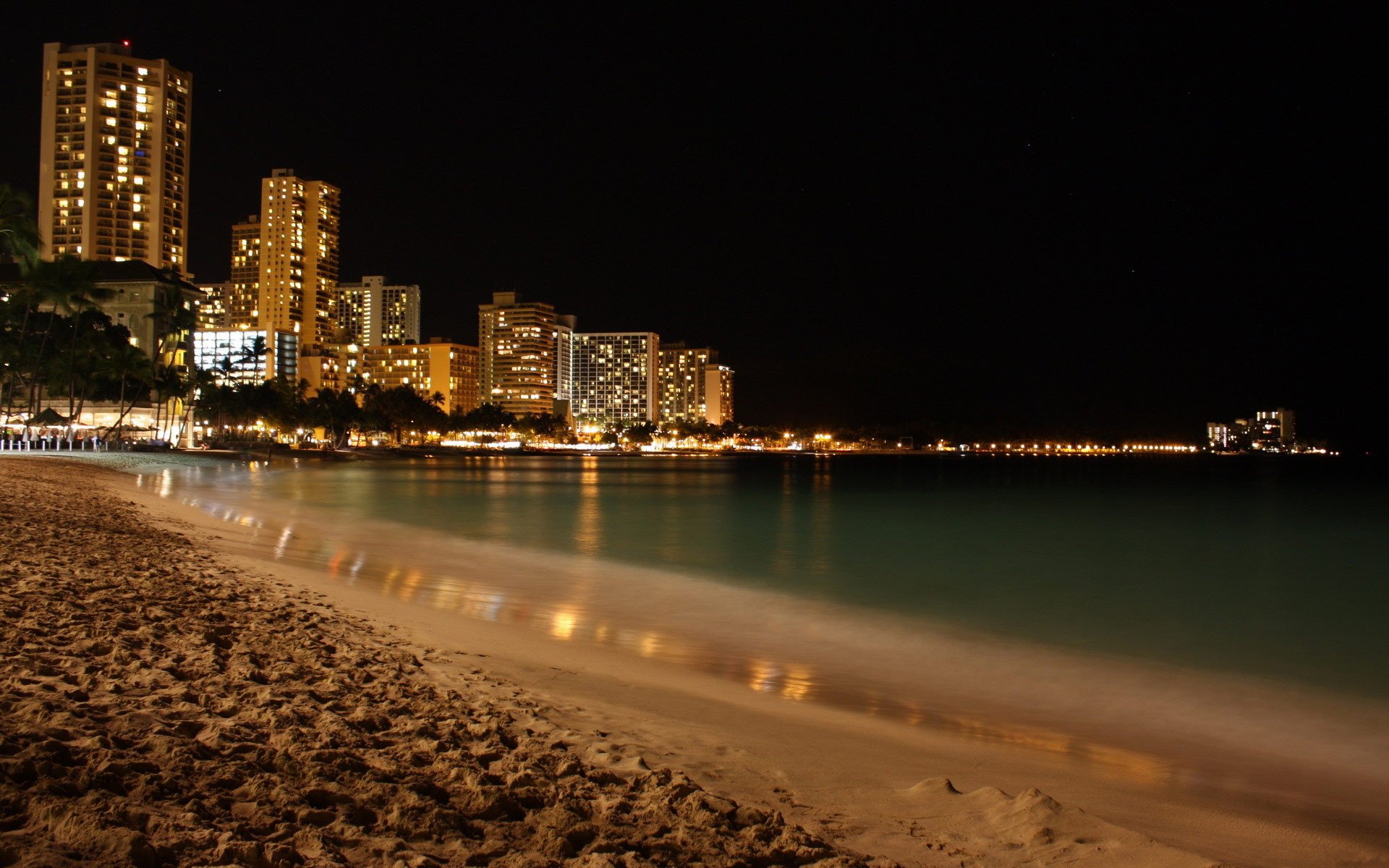 HD photography wallpaper capturing a serene nighttime coastline with city lights reflecting on the calm water and sandy beach in the foreground.