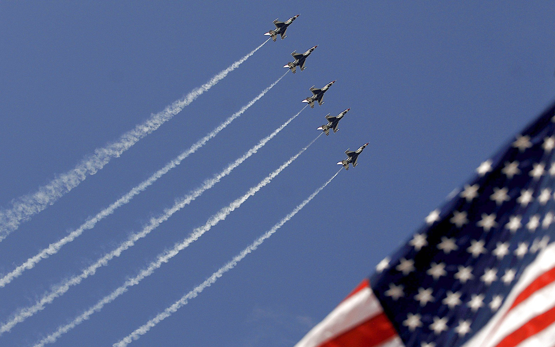 HD PC desktop wallpaper showing military jets in tight formation at an air show, trailing white smoke across a clear blue sky with a waving American flag in the corner.