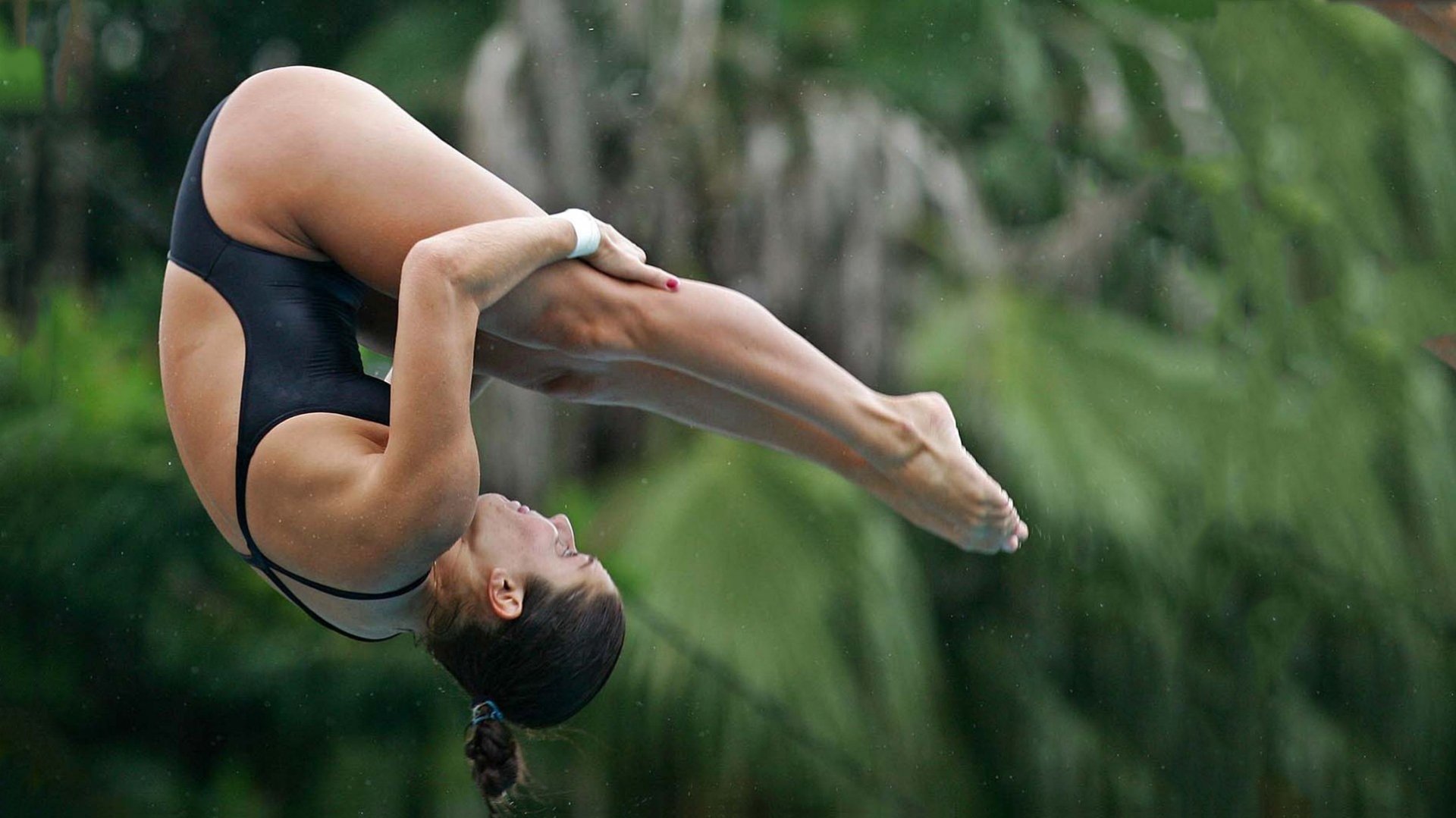 Athletic diver curled in a tight mid-tuck above blurred tropical foliage, dynamic sports HD PC desktop wallpaper and background.
