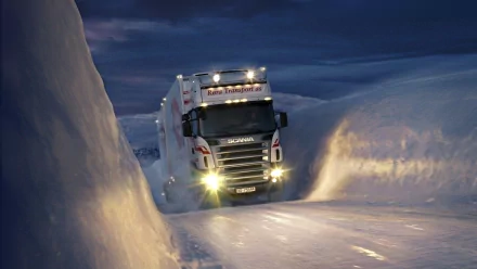 A powerful truck navigates a narrow path through deep snow, illuminated by its bright headlights against a twilight sky, making for a striking HD desktop wallpaper.