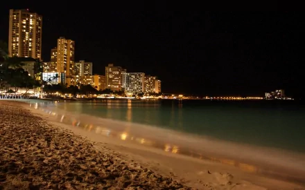 HD photography wallpaper capturing a serene nighttime coastline with city lights reflecting on the calm water and sandy beach in the foreground.