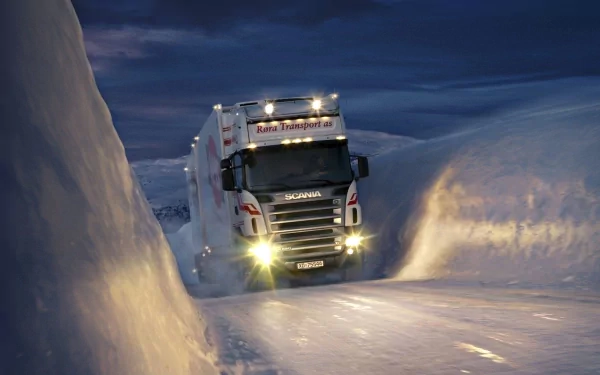 A powerful truck navigates a narrow path through deep snow, illuminated by its bright headlights against a twilight sky, making for a striking HD desktop wallpaper.