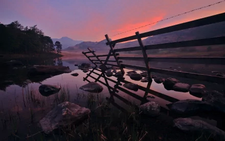 HD wallpaper depicting a partially collapsed wooden fence stretching into a serene lake, reflecting a vibrant sunset sky. Rocky terrain and distant trees line the tranquil water's edge.