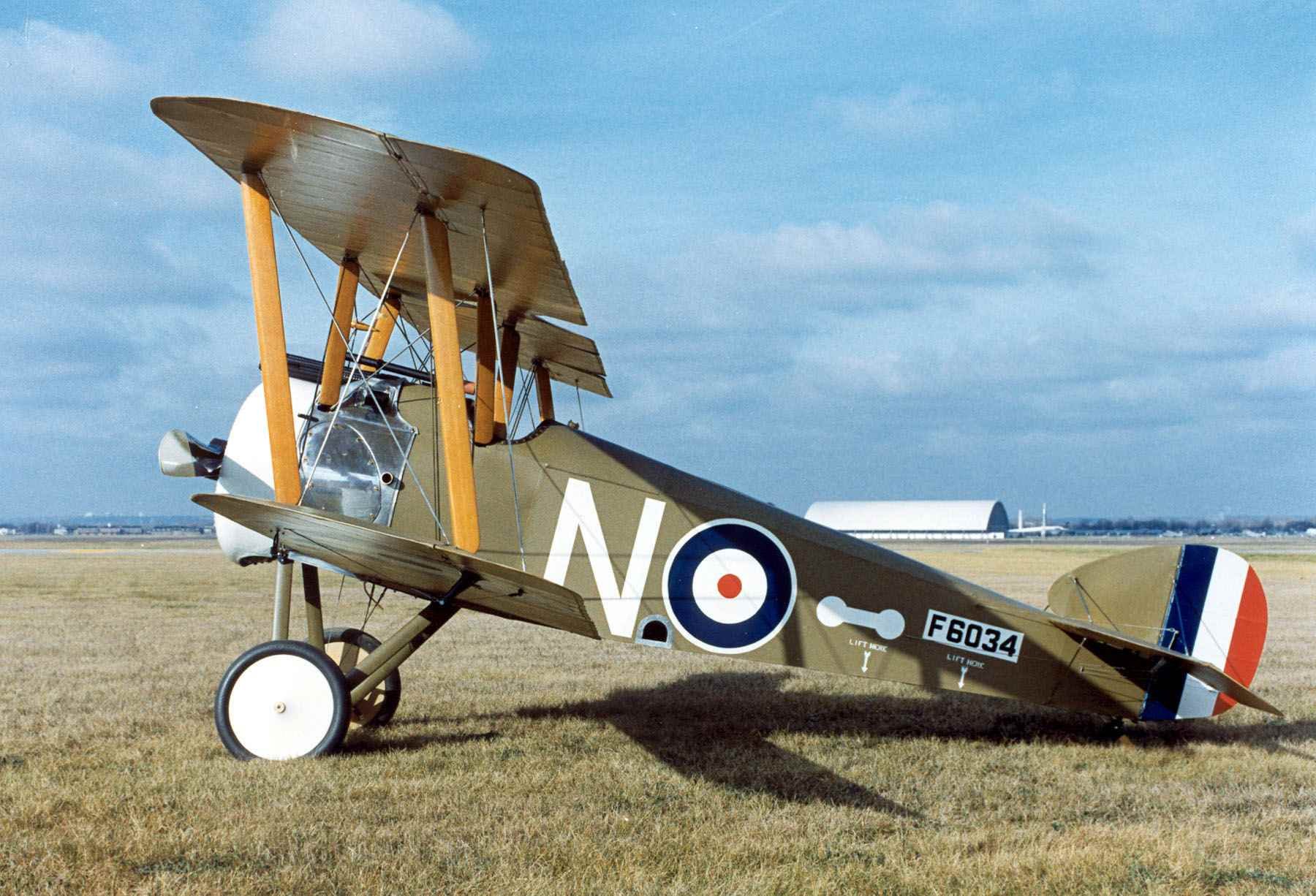 HD desktop wallpaper showing a vintage military biplane with roundels and tail stripes, set on a grassy field under a clear blue sky.
