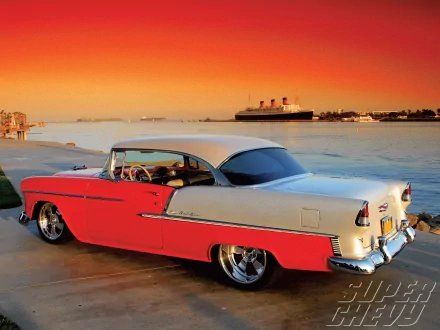 A vibrant sunset backdrop features a classic 1955 Chevy Bel Air parked by the water, showcasing its sleek lines and striking two-tone coloring against a serene landscape.