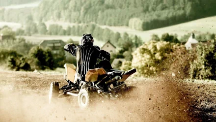 Rear view of a rider on an ATV kicking up dust on a dirt trail with rolling green hills beyond — 2K Quad HD PC desktop wallpaper.