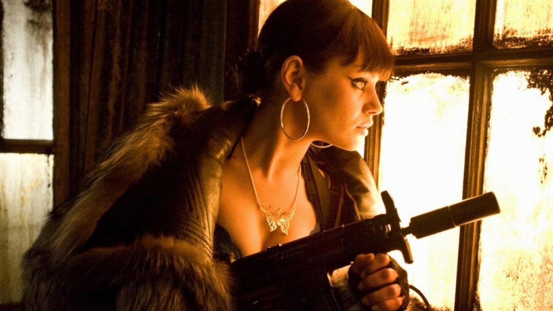 HD PC desktop wallpaper featuring celebrity Mila Kunis in a dramatic scene, holding a firearm with warm lighting from a window behind her.
