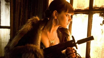 HD PC desktop wallpaper featuring celebrity Mila Kunis in a dramatic scene, holding a firearm with warm lighting from a window behind her.