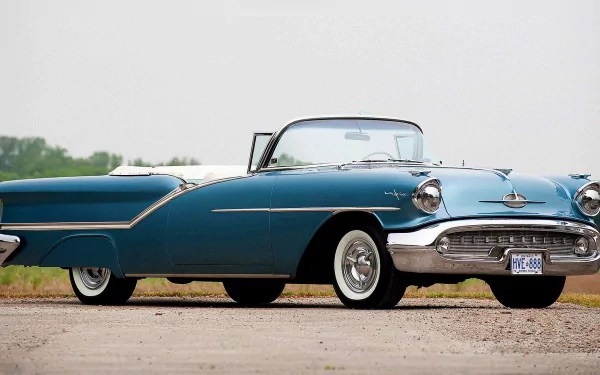 HD desktop wallpaper featuring a vintage blue Oldsmobile convertible parked on a dirt road with a clear sky background.