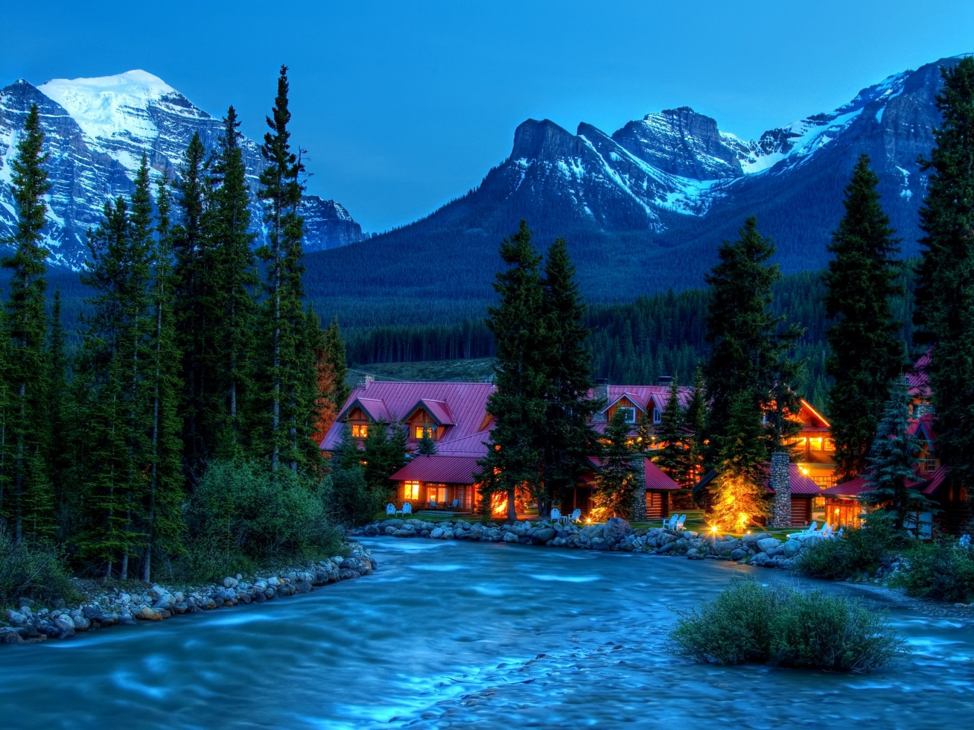 A serene landscape featuring a river flowing beside a house, set against majestic mountains and lush trees, captured at dusk for a stunning HD wallpaper.