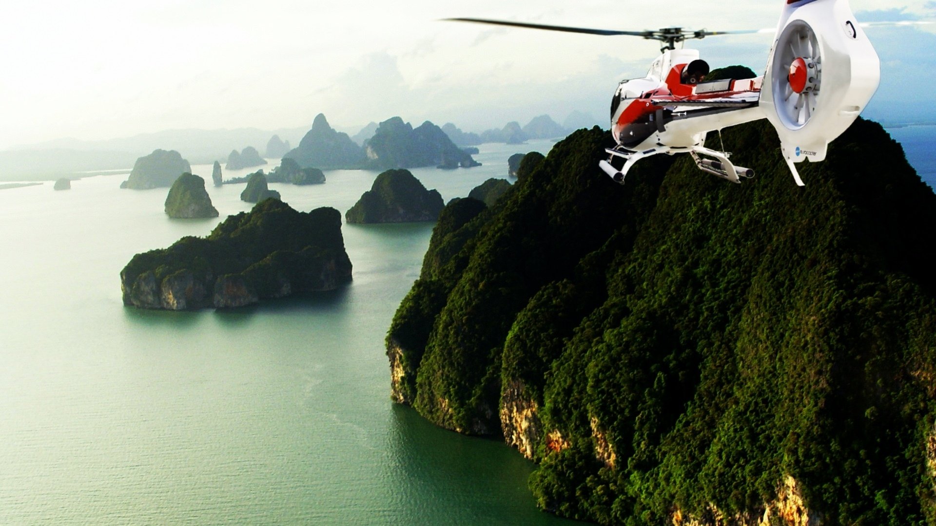 HD PC desktop wallpaper and background showing a helicopter vehicle flying past green cliffs and limestone islands over a calm turquoise sea.