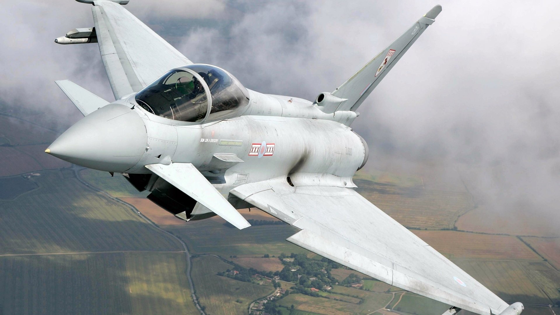 HD PC desktop wallpaper showing a military Eurofighter Typhoon soaring over patchwork countryside beneath a cloudy sky.
