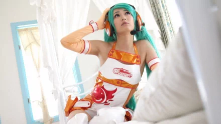 HD desktop wallpaper featuring a woman cosplaying as Hatsune Miku, dressed in an elaborate outfit with a green wig, seated indoors with a white and blue background.