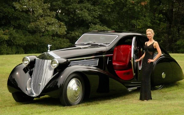 A stylish woman stands beside a vintage black car with a sleek design, set against a lush green backdrop. This HD image captures the elegance of girls and cars.