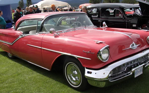 HD desktop wallpaper featuring a classic red Oldsmobile parked on grass at a car show, surrounded by other vintage vehicles and attendees.