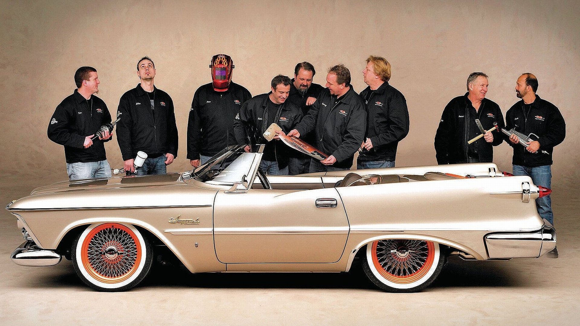 HD PC desktop wallpaper showing a beige vintage Chrysler convertible with wire wheels against a neutral backdrop, a group of men in black jackets standing and chatting behind the car.