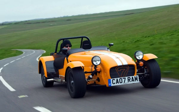 HD PC desktop wallpaper: yellow Caterham open-top sports car with white stripes speeding along a winding country road against green hills.
