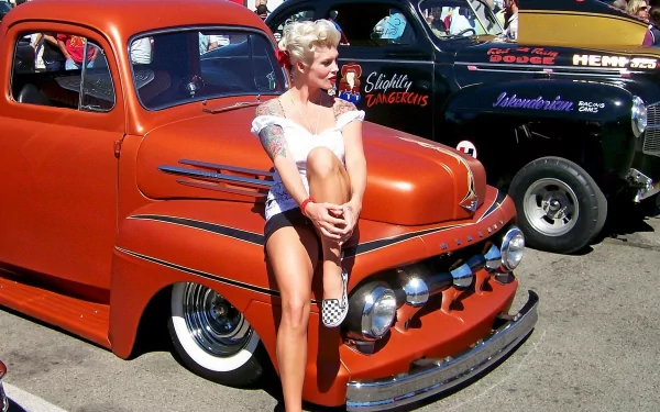 HD desktop wallpaper featuring a woman with vintage hairstyle sitting on the front bumper of a bright orange classic car at an outdoor car show.
