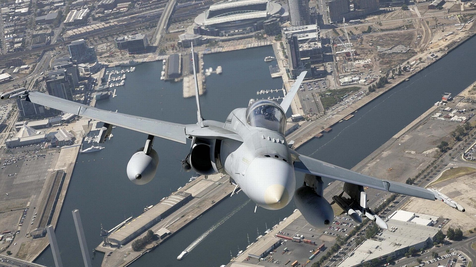 Military McDonnell Douglas F/A-18 Hornet jet soaring above a harbor cityscape, captured in clear detail as an HD PC desktop wallpaper and background.