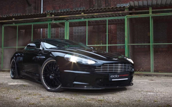 HD desktop wallpaper featuring a sleek black Aston Martin DBS parked in front of a rustic green metal fence and brick wall.
