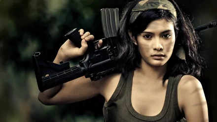 A confident woman holding a gun, wearing a tank top and headband, set against a blurred outdoor background. This HD wallpaper captures strength and determination.