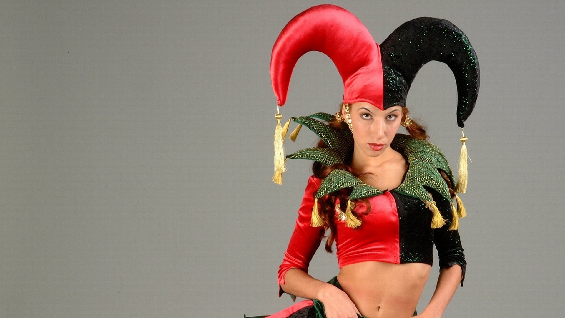 HD PC desktop wallpaper of a woman in jester cosplay: red-and-black horned hat, green ruffled collar, and cropped red-and-black costume against a gray background.