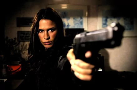 HD PC desktop wallpaper featuring Rhona Mitra from the movie Skinwalkers, aiming a handgun with an intense expression in a dimly lit setting.