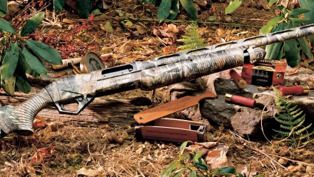 HD desktop wallpaper featuring a camouflaged man-made shotgun resting on forest ground with scattered shotgun shells and foliage background.