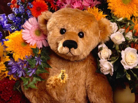 A high-definition desktop wallpaper featuring a stuffed teddy bear surrounded by an array of vibrant flowers, including roses, daisies, and lilies.