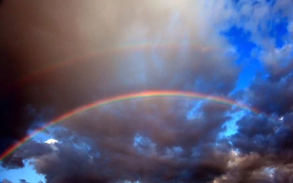 HD desktop wallpaper featuring a vivid double rainbow arching across a dramatic sky filled with dark clouds and patches of bright blue.