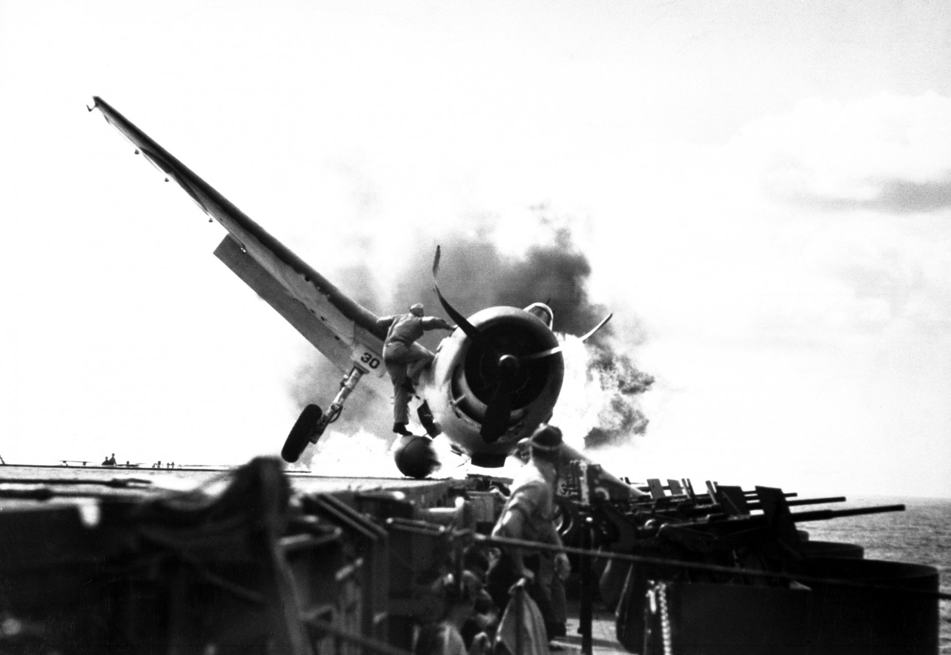 Black and white HD wallpaper of a military Grumman F6F Hellcat in action, with smoke billowing around the aircraft, captured on a carrier deck.