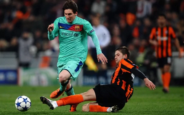 HD desktop wallpaper featuring Lionel Messi in action during a football match, wearing a turquoise Barcelona kit and evading a slide tackle from an opponent in an orange and black jersey.