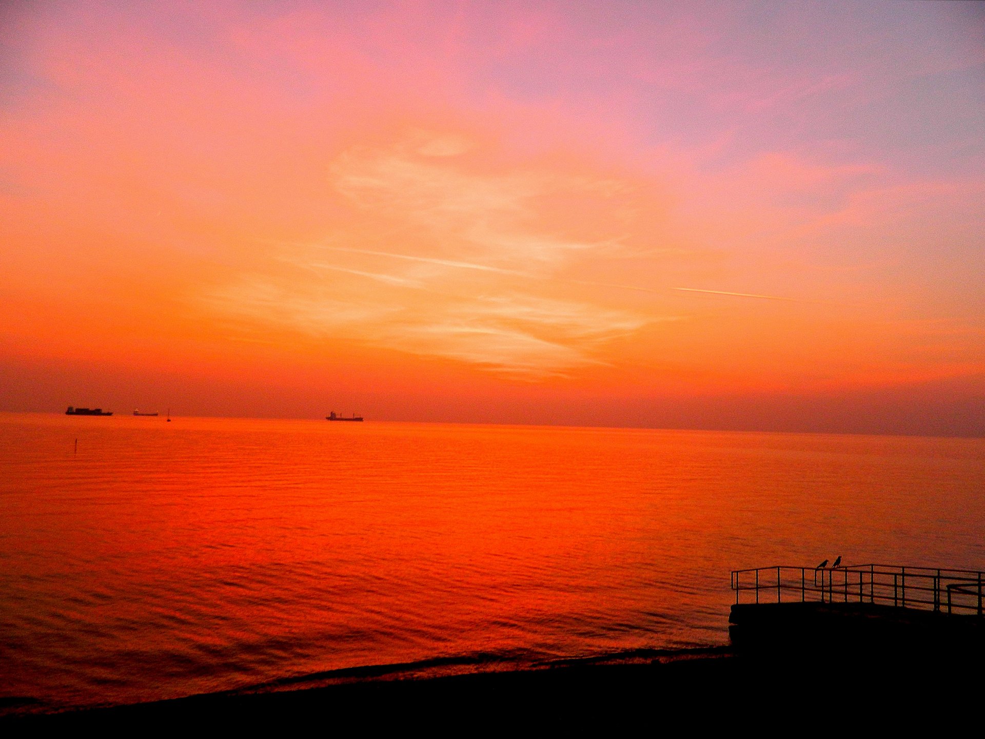 4K Ultra HD nature photography capturing a vibrant orange sunset over a calm sea, with a silhouetted railing in the foreground and ships on the horizon.