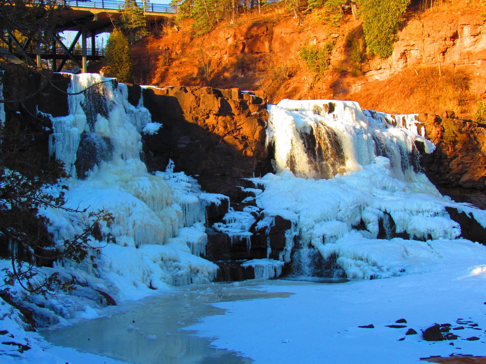 HD PC desktop wallpaper and background: scenic nature view of a partially frozen waterfall cascading under sunlit red cliffs, blue ice and snow along the icy pool.