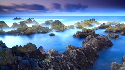 A serene seascape captures gentle waves lapping against rugged rocks, with a tranquil blue hue and soft clouds above, creating a stunning nature-inspired HD desktop wallpaper.