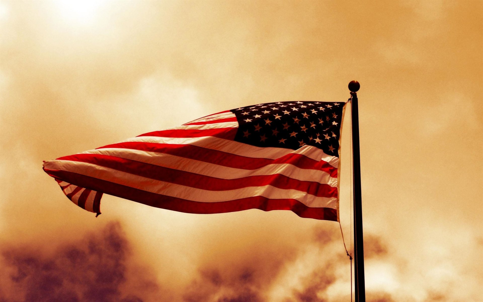 A high-definition desktop wallpaper featuring the American flag waving against a dramatic sky. The colors and textures create a striking visual effect.