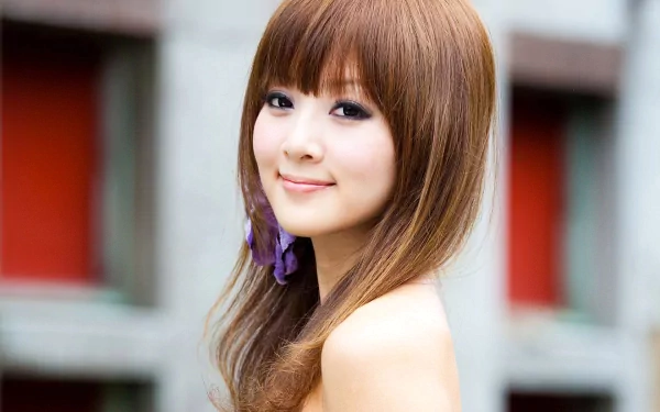 HD desktop wallpaper and background featuring an Asian woman, Mikako Zhang Kaijie, with long brown hair, smiling gently in an outdoor setting with blurred architectural elements in the background.