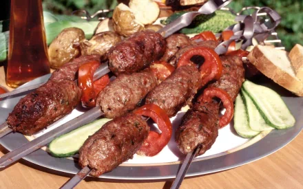 HD PC desktop wallpaper showing a plate of barbecue skewers with grilled meat, tomato slices, baked potatoes, cucumber, and bread in an outdoor setting.