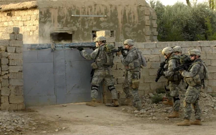 HD wallpaper showing U.S. Army Infantry soldiers in full military gear during a war operation, positioned beside a weathered brick structure.