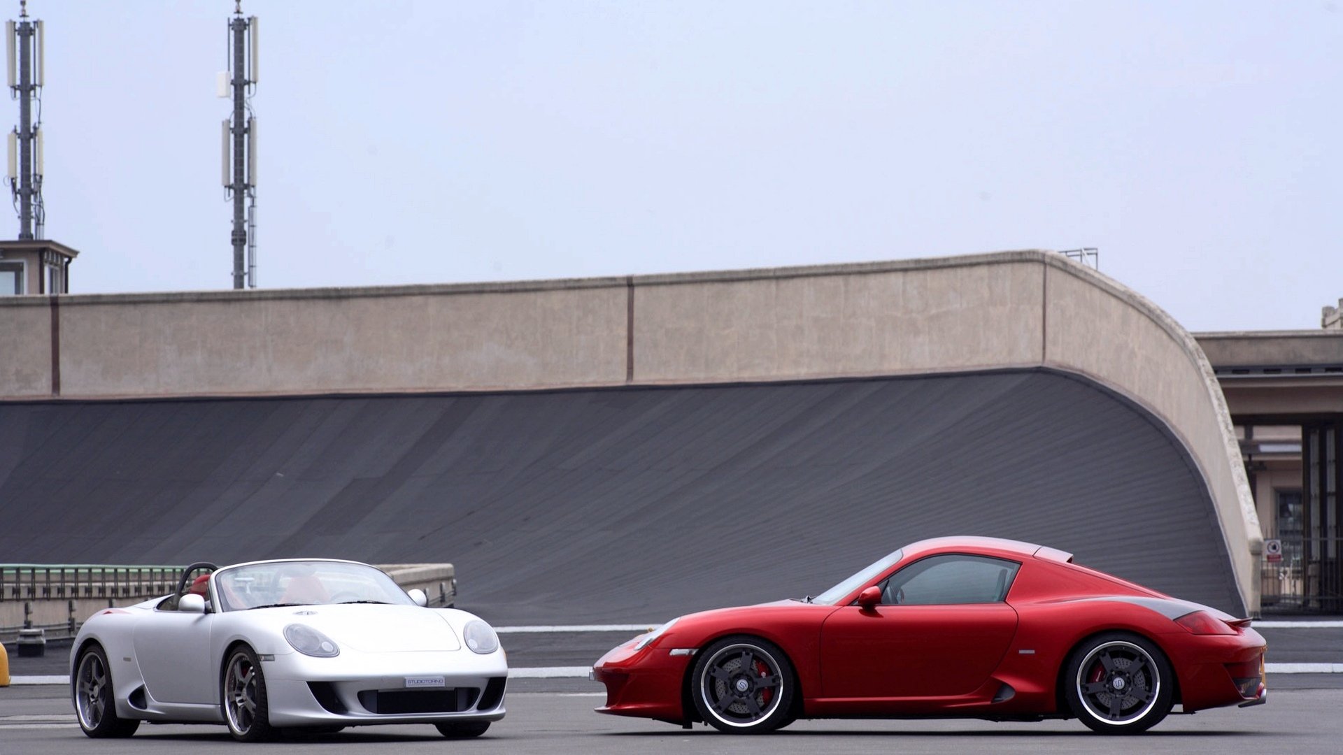 HD PC desktop wallpaper of two sports cars—a red coupe and a silver convertible—parked side by side on a racetrack in front of a curved concrete wall.